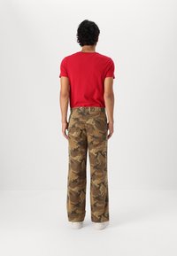 Rode t-shirt met korte mouwen en ronde hals, gecombineerd met loszittende, tan camouflagemotief broeken en witte sneakers, gezien vanaf de achterkant.