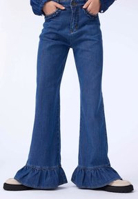 Blå denim bukser med hjælpet nederdel, høy midje, lommer og kontrastsømm, kombinert med kremfargede og svarte plattformsko.