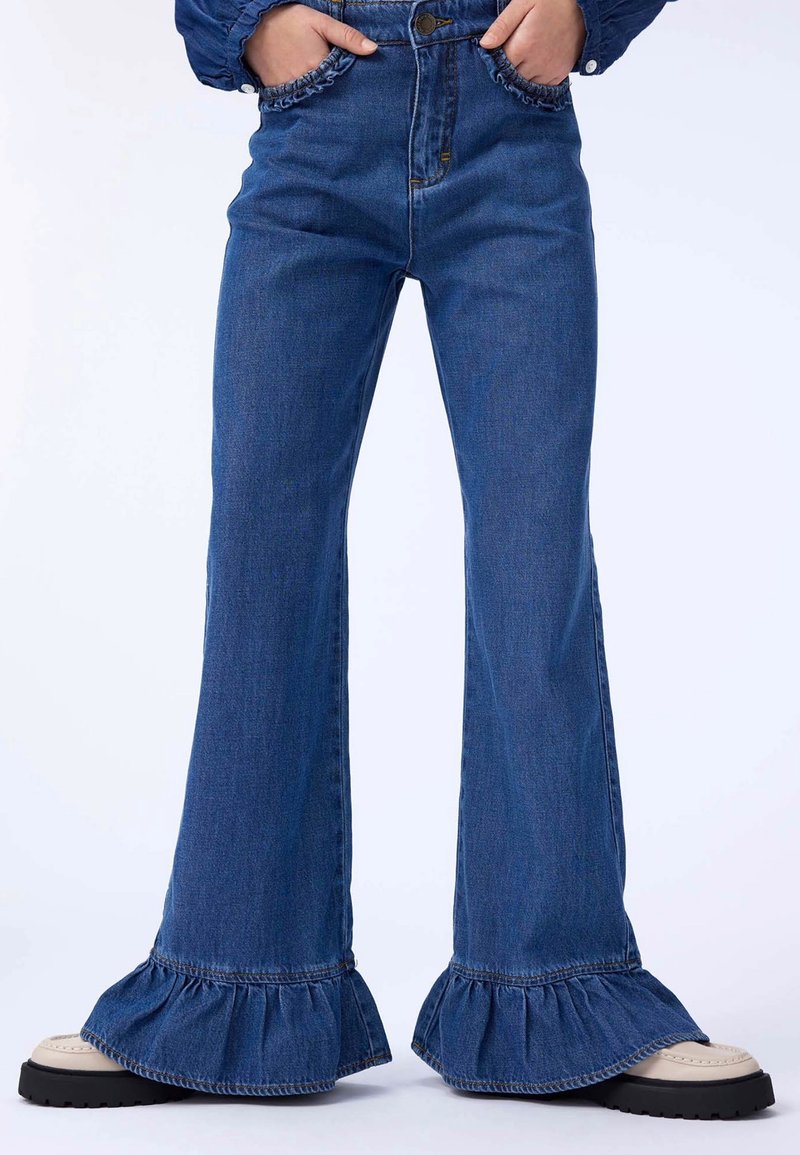 Blå denim bukser med hjælpet nederdel, høy midje, lommer og kontrastsømm, kombinert med kremfargede og svarte plattformsko.