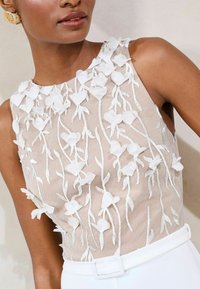 Top sin mangas de tela beige transparente adornado con apliques florales blancos y bordado texturizado, con un cuello alto y una cinturilla.