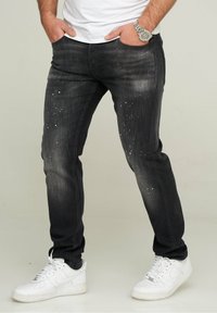 Vaqueros de mezclilla negros con un ajuste slim, detalles desgastados y salpicaduras de pintura blanca. Combinados con zapatillas deportivas blancas.