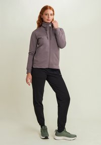 Giacca grigia con zip e colletto alto, con tasche frontali, abbinata a pantaloni jogger neri e sneaker verdi. Tessuto liscio con dettagli a motivo sottile.