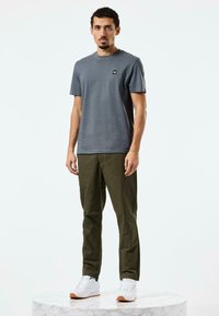 Grå bomulls t-skjorte med en liten logo, kombinert med olivengrønne bukser og hvite sneakers. Modellen står på en marmor pedestal.