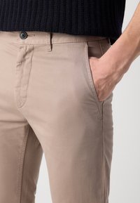 Chinos beige en tissu léger avec une coupe droite, devant plat et poches latérales. Présentent une fermeture à bouton et des détails de couture discrets.