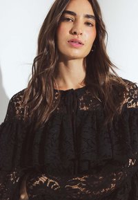 Juodas nėrinių topas su raukšlėtomis rankovėmis, puikuojantis tekstūruotu gėlių raštu ir užsegimu prie kaklo. Modelis pozuoja su natūraliu makiažu.