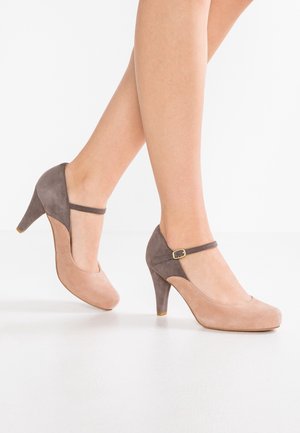 Jambes portant des talons hauts Mary Jane en daim beige et marron avec des brides à la cheville, marchant sur un sol blanc lisse sur un fond blanc uni.