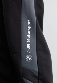 Fekete pulóver szürke részletekkel, a "Motorsport" felirattal és a BMW logóval az ujján. Az anyag lágy és textúrált felületűnek tűnik.