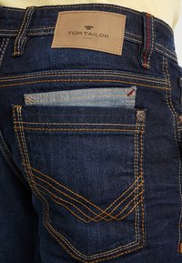 Donkerdenim jeans met oranje stiksels, voorzien van een leren label met de tekst "TOM TAILOR," een klein zakje dat eruitsteekt, en unieke stiksels op de achterzak.