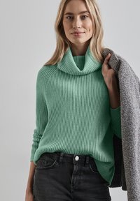 Myntgrön stickad tröja med stor turtleneck och ribbad textur, i kombination med mörka jeans och håller en grå texturerad jacka.