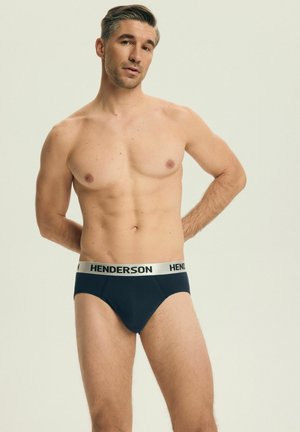 Mężczyzna bez koszulki o krótkich ciemnych włosach, ubrany w granatowe bokserki z białym pasem w talii z napisem "HENDERSON", stojący z rękami za plecami.