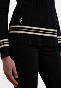 Navy cable-knit sweater with ribbed cuffs and hem, featuring cream stripes at the bottom, and a logo detail on the front.