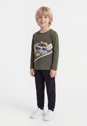 Jeune garçon debout et souriant, portant un t-shirt à manches longues vert olive avec des motifs de véhicules de construction, un pantalon noir et des baskets blanches.