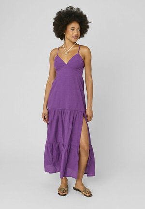 Femme avec coiffure afro portant une robe maxi violette sans manches avec une fente latérale, collier soleil en or et sandales fleuries, souriante et regardant de côté.