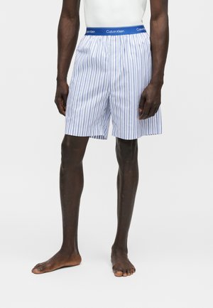 Hombre vistiendo bóxers Calvin Klein a rayas verticales blancas y azules con cintura elástica azul con marca, de pie descalzo sobre fondo blanco.