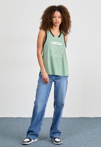 Grön maskinvävd basketlinne med svarta accenter och vit text, kombinerat med ljusblå vida jeans och svart-vita sneakers.