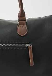 Bolso negro texturizado con acentos de cuero marrón, que cuenta con un bolsillo con cremallera, un accesorio de asa redonda y una superficie con patrón.