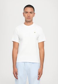 White cotton t-shirt with short sleeves, crew neckline, and small embroidered detail on the chest. Model wears striped blue pajama pants.