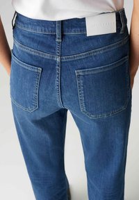 Blauwe spijkerbroek met een hoge taille, vijf zakken en een wit leren logopatch op de achterkant. De stof heeft een lichte textuur.