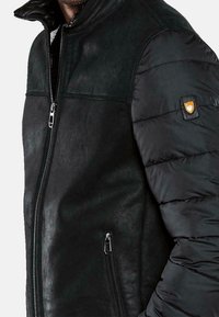 Musta takki, jossa on pehmustettu muotoilu ja sileä, matta pinta vartalossa sekä tilkkutekstuuri hihoissa. Vetoketju ja logoyksityiskohta.