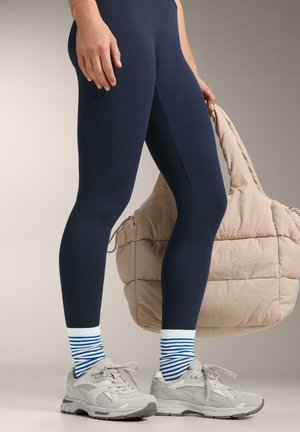 Personne portant des leggings bleu marine, des chaussettes blanches à rayures bleues, des baskets grises, tenant un grand sac matelassé beige par les poignées.