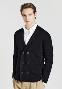 Schwarzer doppelbreasted Cardigan aus gestricktem Stoff mit großen blauen Knöpfen und einem tiefen V-Ausschnitt, kombiniert mit einem weißen Hemd.