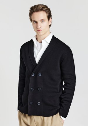 Black double-breasted cardigan made of knitted fabric, featuring large blue buttons and a deep V-neck design, paired with a white shirt.