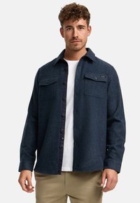 Hombre con camisa de lana azul oscuro abotonada sobre camiseta blanca y pantalones beige, de pie contra un fondo claro y liso.