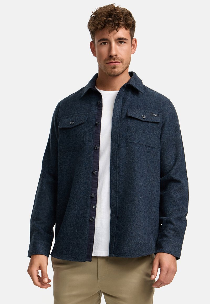 Hombre con camisa de lana azul oscuro abotonada sobre camiseta blanca y pantalones beige, de pie contra un fondo claro y liso.