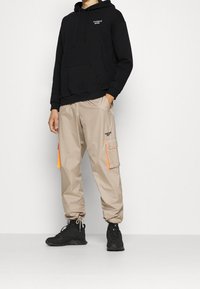Felpa nera con cappuccio e tasca frontale, abbinata a pantaloni cargo beige con accenti arancioni e tasche laterali, indossati con calzature nere.