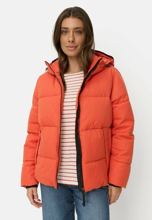 Frau trägt eine orangefarbene Steppjacke mit Kapuze, ein weiß-orange gestreiftes Oberteil und blaue Jeans, stehend vor einem weißen Hintergrund.