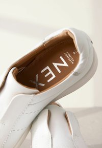 Hvide slip-on sko med tekstureret læderoverdel, rund tå og kontrasterende tan indersål med sølv branding. Minimalistisk design med syede detaljer.