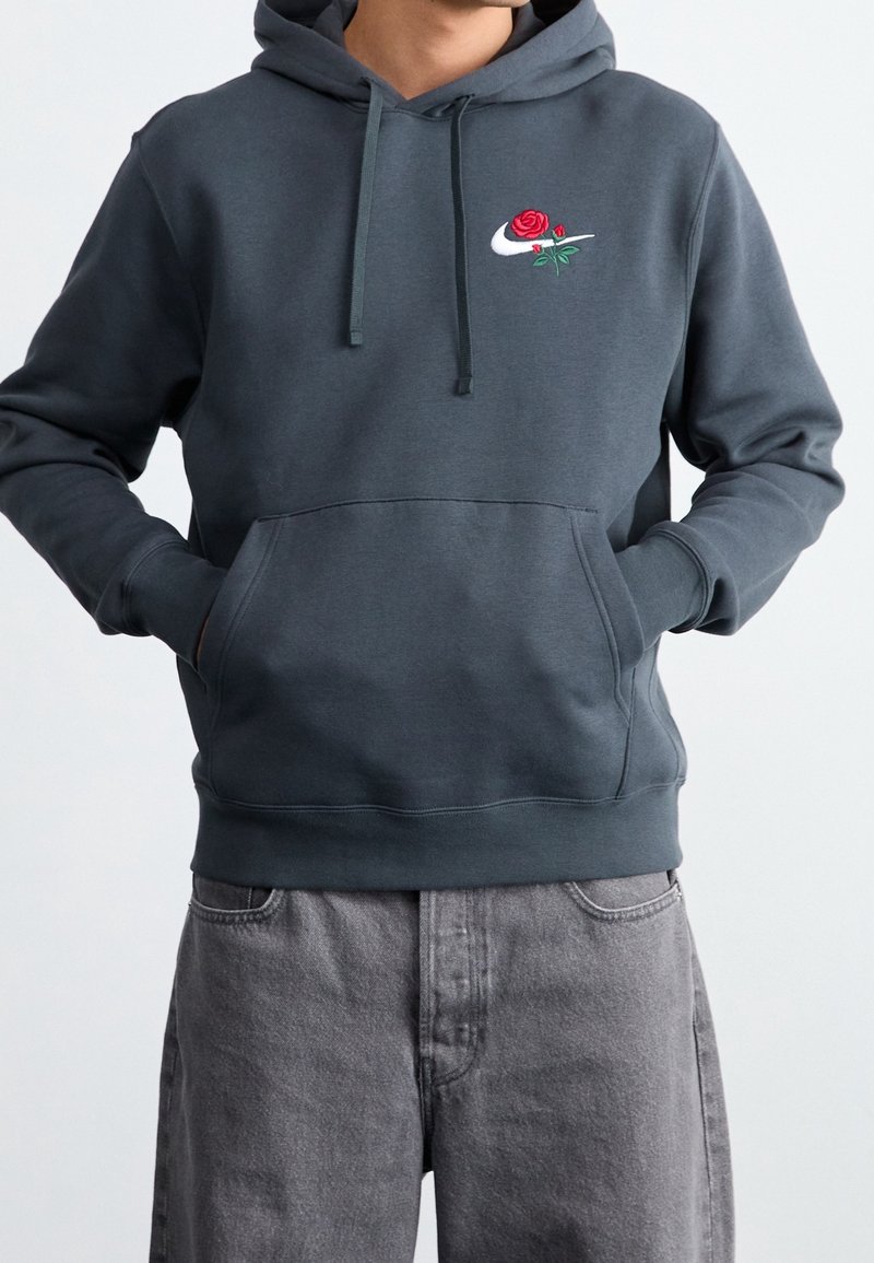 Mörkgrå hoodie med en broderad ros och Nike-logotyp på bröstet. Har en kenguruficka och justerbar dragkedja på huvan.