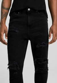 Vaqueros de mezclilla negra con un corte slim, que presentan detalles desgastados y parches deshilachados. Cierre de botón estándar y diseño de cinco bolsillos.