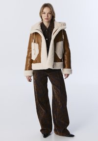 Bruine leren jas met een crèmekleurige schapenvachtvoering, oversized capuchon en twee grote zakken; gecombineerd met wijde, gestreepte broeken.