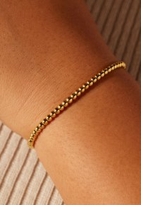 Gouden armband met kleine ronde kralen gedragen om een pols tegen een neutrale, gestructureerde stoffen achtergrond.