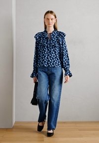 Blusa blu marina con motivi a cuore azzurri, maniche a volants e scollatura con lacci, abbinata a jeans larghi azzurri e scarpe nere.