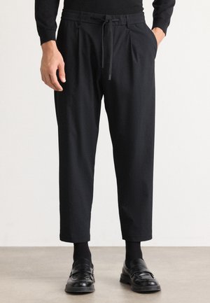 Black trousers feature a relaxed fit with a textured fabric, pleated front, elastic waistband with drawstring, and tapered legs.