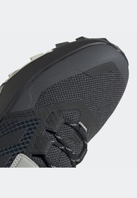 Chaussure de sport grise dotée d'une tige en mesh texturé, d'accents noirs renforcés et d'une semelle extérieure en caoutchouc robuste avec des crampons multidirectionnels.