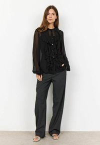 Black blouse with ruffled front detail, sheer long sleeves, and button closures, paired with loose gray trousers and silver sandals.