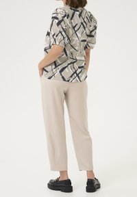 Kvinde iført beige bukser, sorte loafers og en kortærmet top med sorte og beige abstrakte penselstræk mønstre, hænderne i lommerne.