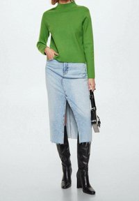 Pull vert en maille à col montant, associé à une jupe en jean bleu clair avec une fente sur le devant. Bottes noires à talons hauteur genou. Sac à main noir.