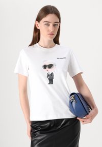 Weißes Baumwoll-T-Shirt mit einem Cartoon-Graphic eines Figuren in einem schwarzen Anzug und Sonnenbrille, mit "KARL LAGERFELD" darüber gedruckt.