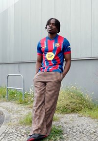 FC Barcelona marškiniai mėlynos ir raudonos spalvos su dryžiais, derinami su rudos spalvos languotomis plačiomis kelnėmis ir juodais batais. Neutralus fonas su žaluma.