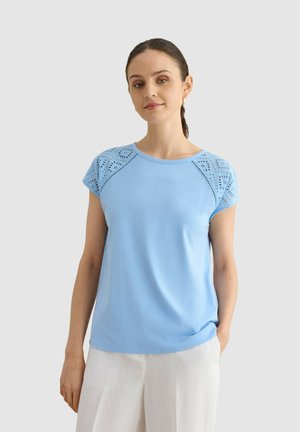Mujer con cabello oscuro, que lleva una camiseta azul clara con mangas de encaje ojete y pantalones blancos, de pie contra un fondo claro y liso.