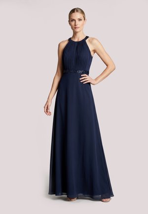 Vrouw staat met hand op heup, draagt een mouwloze marineblauwe geplooide halterjurk tot op de vloer met een tailleband met kralen.