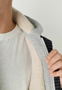 Kapuzensjacke mit grauem Außenmaterial und weichem, beigem Fleecefutter; verfügt über einen durchgehenden Reißverschluss und ein strukturiertes, dunkles Material am Kragen.