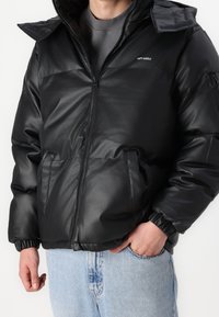 Svart puffjacka med dragkedja fram, elastiska ärmmuddar och sidofickor. Har en logotyp på vänster bröst och en bred huva. Glansigt material.