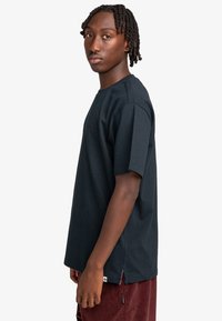 Sort bomulds T-shirt med korte ærmer, rund halsudskæring og sideslids. Har et diskret logo på brystet. Modellen står vendt til siden.