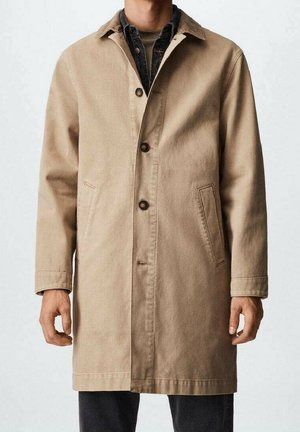 Mężczyzna w beżowym, zapiętym trenczu na ciemnej koszuli i czarnych spodniach, stojący z ramionami swobodnie opuszczonymi wzdłuż ciała.