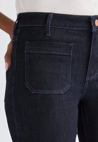 Dark blue denim jeans with back pocket and belt loops, worn by a person with hand resting on hip against plain background.